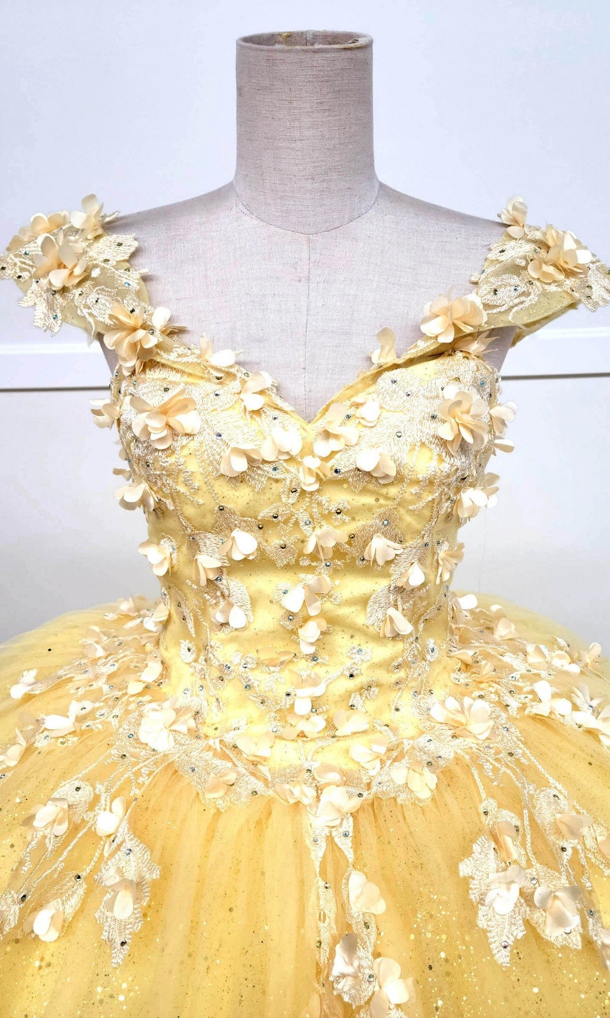The Quinceanera Dress CHF3074 By Chicas is showcased on a mannequin, boasting a vibrant yellow off-shoulder design adorned with intricate lace embroidery. Its voluminous glitter tulle skirt is beautifully embellished with delicate beads and small fabric flowers.